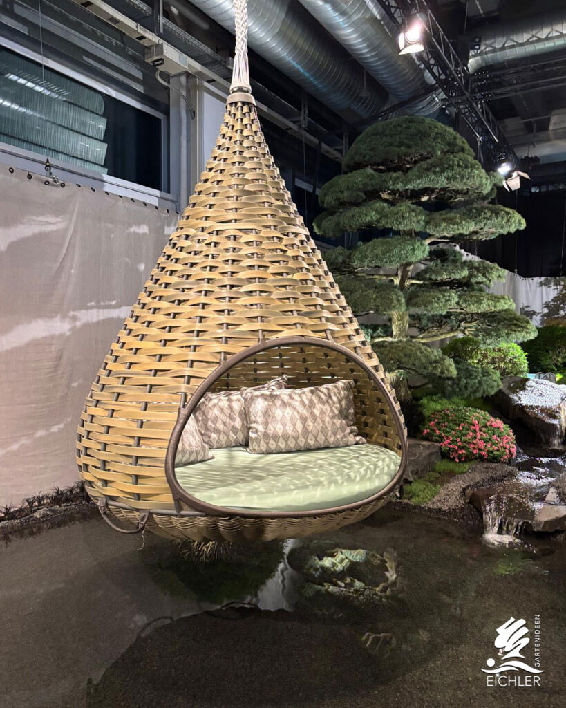 Ein großer, tropfenförmiger Hängesessel aus Geflecht schwebt über einem flachen Wasserbecken, umgeben von Bonsai-artigen Nadelbäumen.