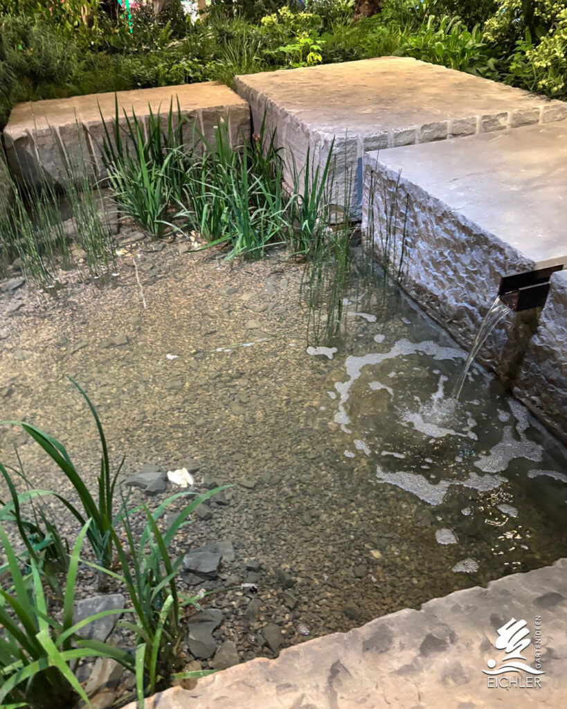 Nahaufnahme eines Wasserbeckens mit grobem Kieselgrund und Gräsern, in das Wasser aus einer massiven Steinmauer fließt.
