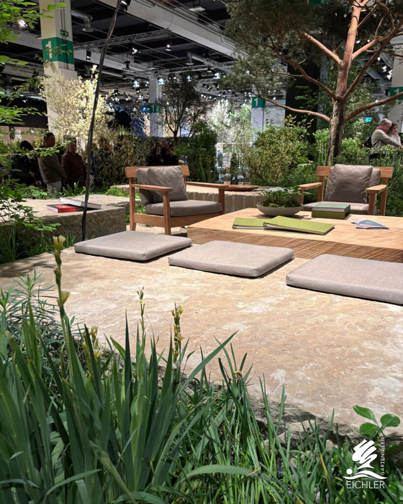 Moderner Loungebereich im Freien mit Holzsesseln, Sitzkissen auf Natursteinplatten und einem flachen Holztisch vor einer grünen Kulisse.