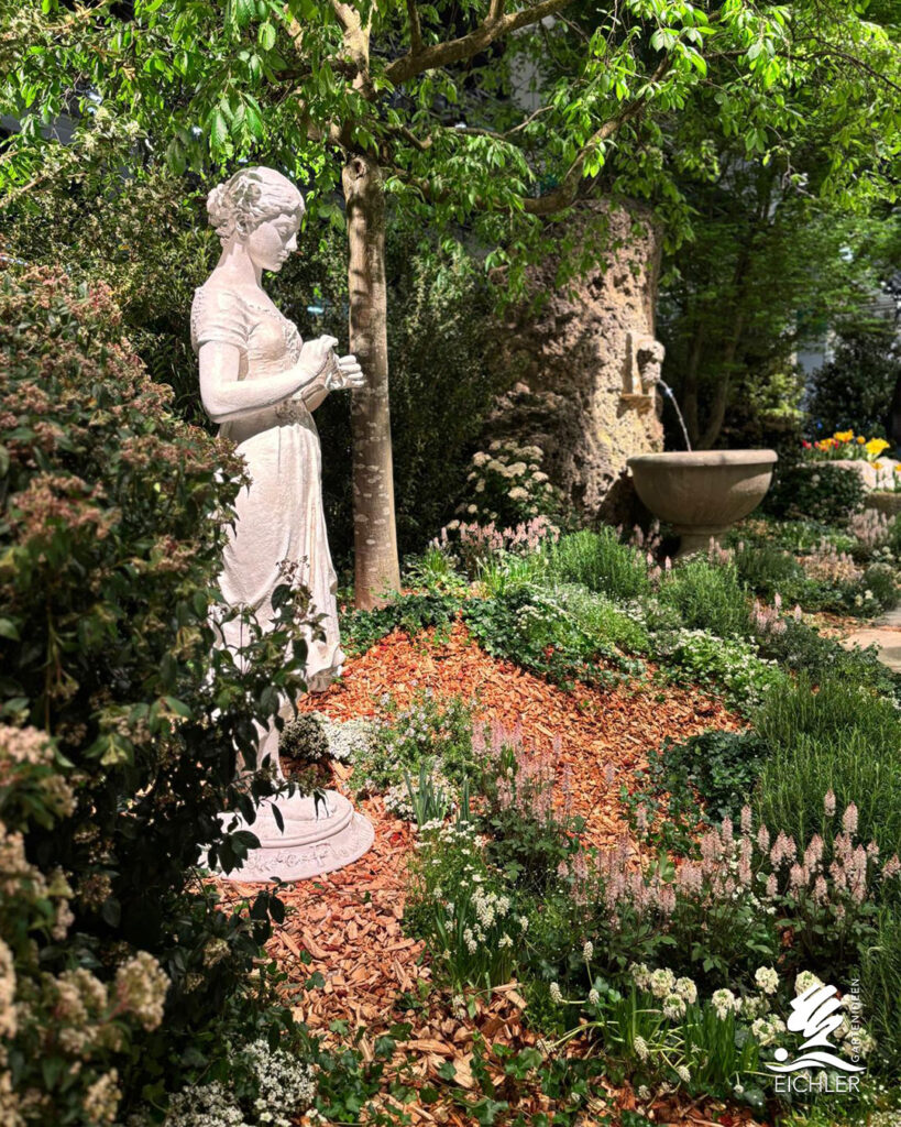 Romantische Gartenecke mit einer weißen Statue einer Frau, umgeben von blühenden Stauden, Rindenmulch und einem Springbrunnen im Hintergrund.