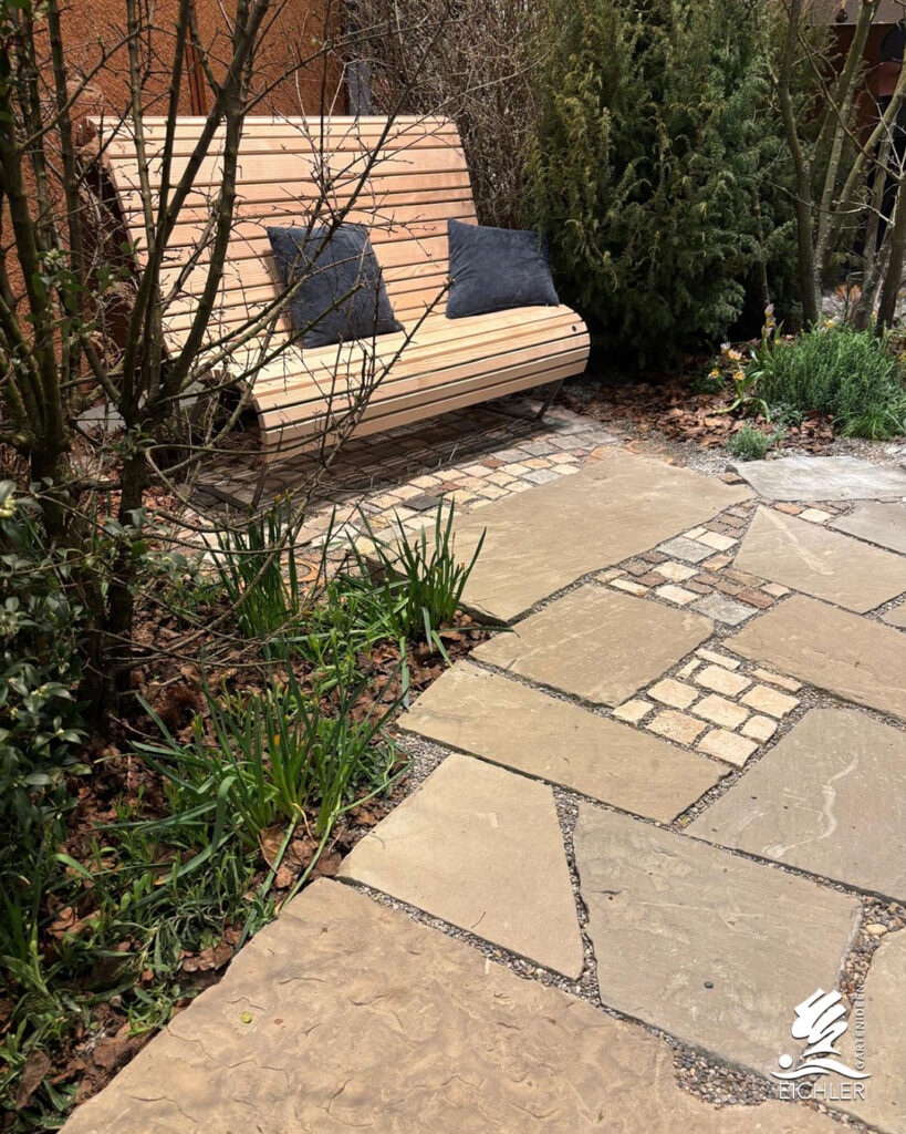 Eine geschwungene Holzbank mit dunklen Kissen auf einer Terrasse aus unregelmäßigen Natursteinplatten und kleinen Pflastersteinen.