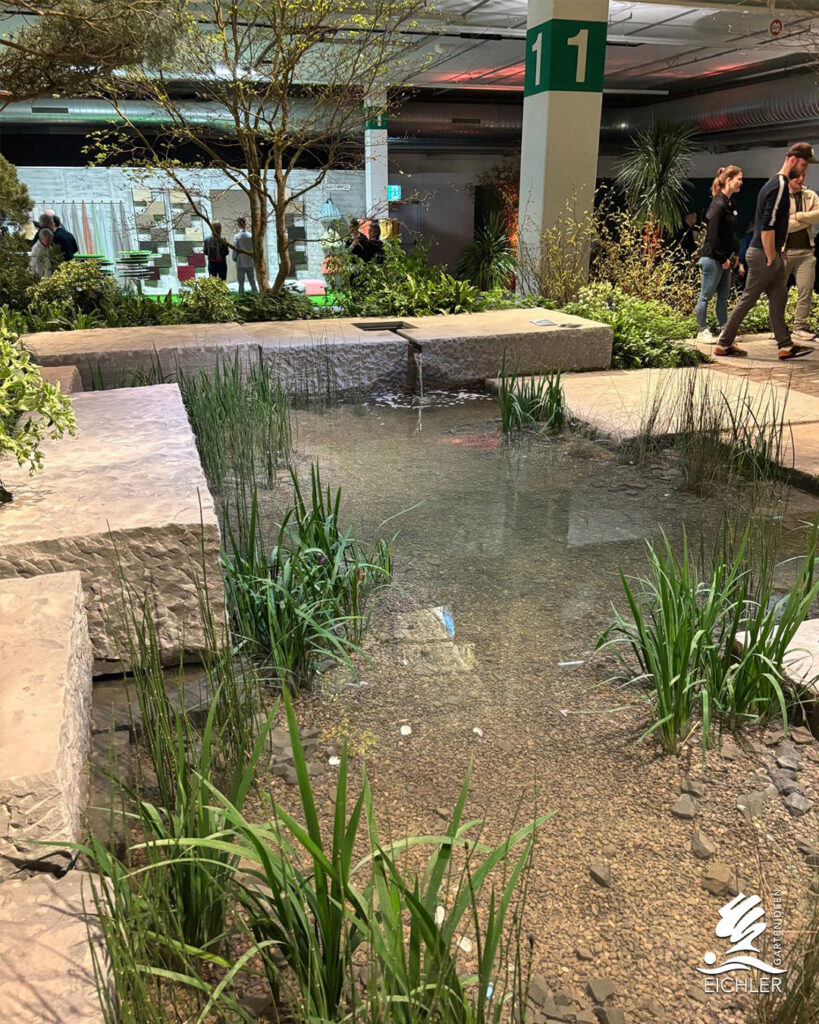 Weitläufige Gartenanlage in einer Messehalle mit einem flachen Teich, hohen Schilfgräsern und großen, rechteckigen Steinelementen.