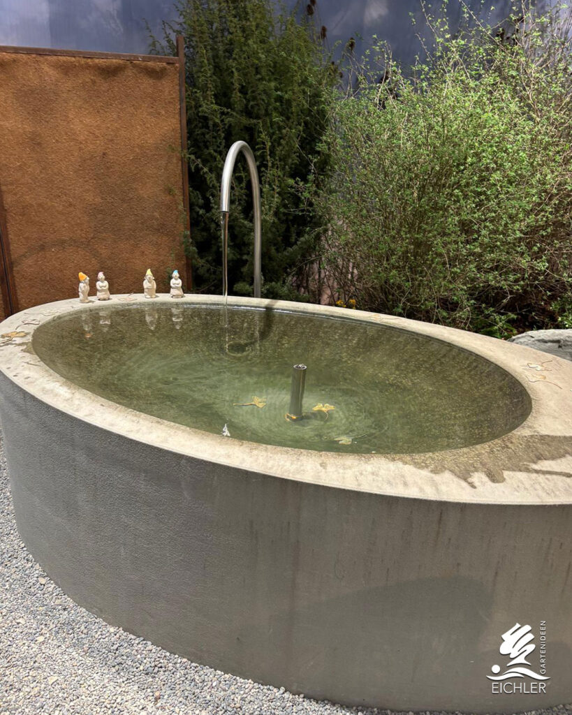 Ovales, monolithisches Betonbecken mit Wasserlauf, kleinen Zwergenfiguren am Rand und Kiesboden im modernen Design.