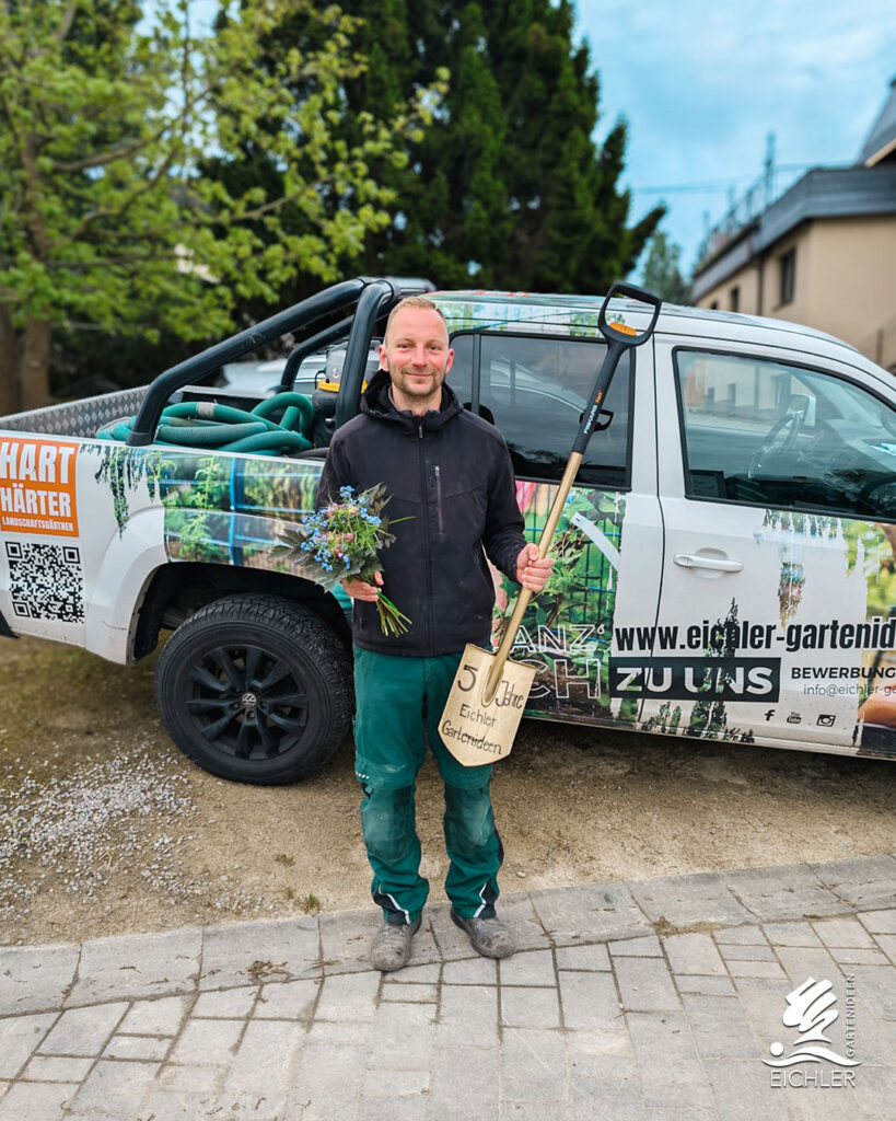 5-Jahre Jubiläum von Mitarbeiter Chris. Er steht mit einem Blumenstrauß und einem goldenen Spaten von Eichler Gartenideen vor einem Betriebswagen im Eichler Design