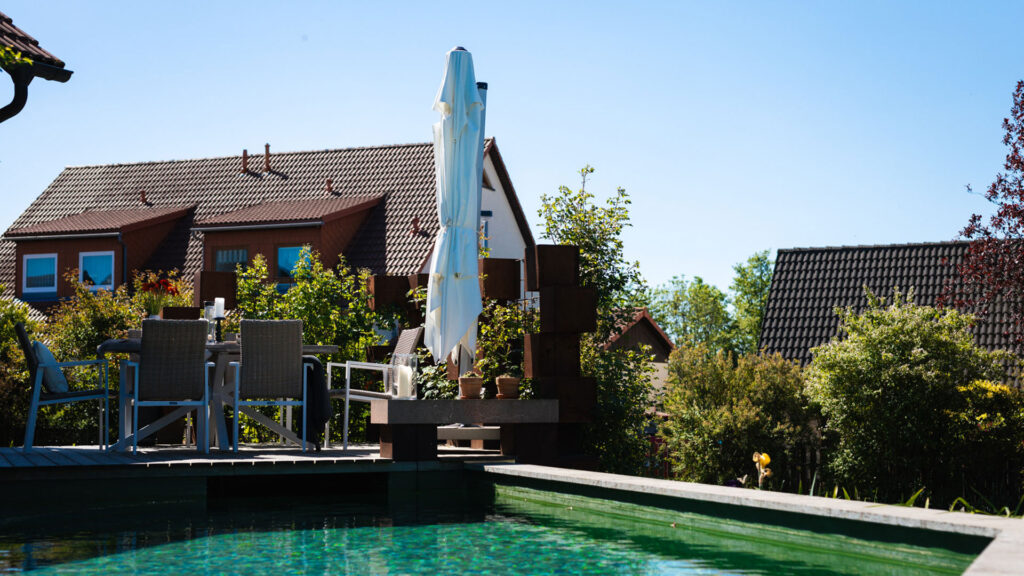 Blick über die Wasseroberfläche eines Pools auf eine Terasse Sitzgruppe im Garten