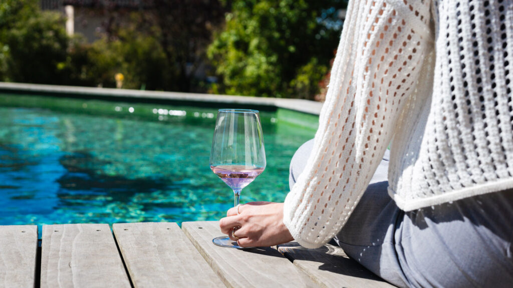 Eine Person sitzt am Poolrand und hält ein Glas Roséwein