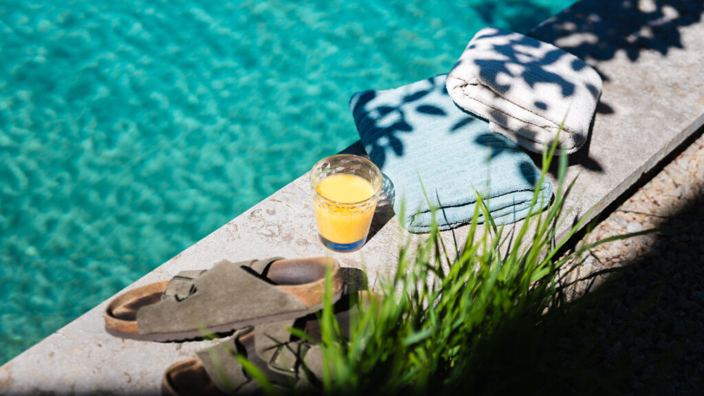 Ein Glas Orangensaft, Sandalen und ein Handtuch liegen am Rand eines Pools