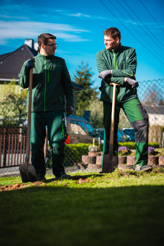 Zwei Landschaftsgärtner in grüner Arbeitskleidung von Eichler Gartenideen stehen entspannt auf einer Rasenfläche, stützen sich auf ihre Spaten und unterhalten sich gut gelaunt bei Sonnenschein.