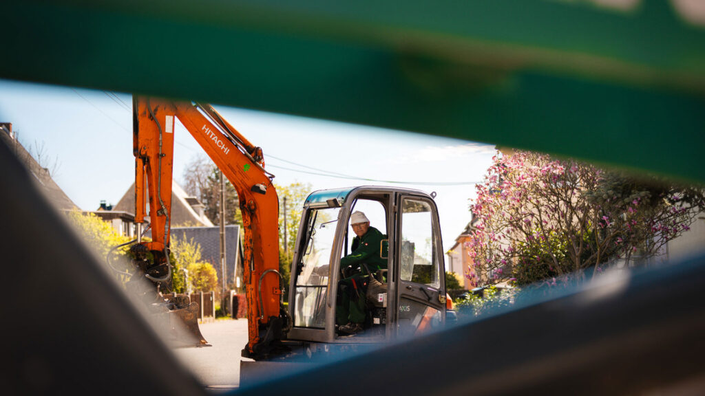 Von hinten fotografiert: Ein Mitarbeiter von Eichler Gartenideen mit weißem Hut dirigiert den Transport eines großen Baumes, der von einer Bagger-Palettengabel am Wurzelballen angehoben wird.