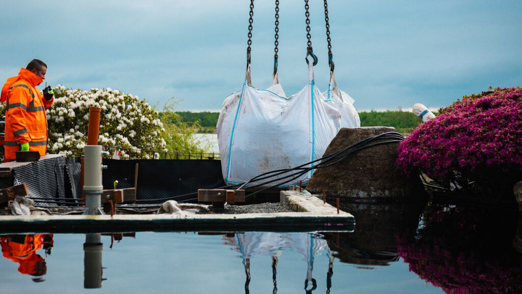 großer, weißer Big Bag mit Schüttgut hängt an den Ketten eines Krans und schwebt über einer Wasserfläche (Teich oder Pool). Ein Arbeiter in orangefarbener Warnjacke beobachtet die Szene am linken Bildrand.