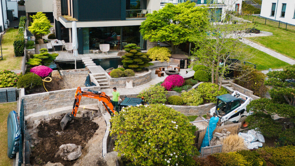 Vogelperspektive auf eine moderne, terrassenförmig angelegte Gartenanlage mit Pool und Designermöbeln. Im Vordergrund hebt ein orangefarbener Minibagger Erde für das neue Baumbeet aus, während ein Gärtner danebensteht.