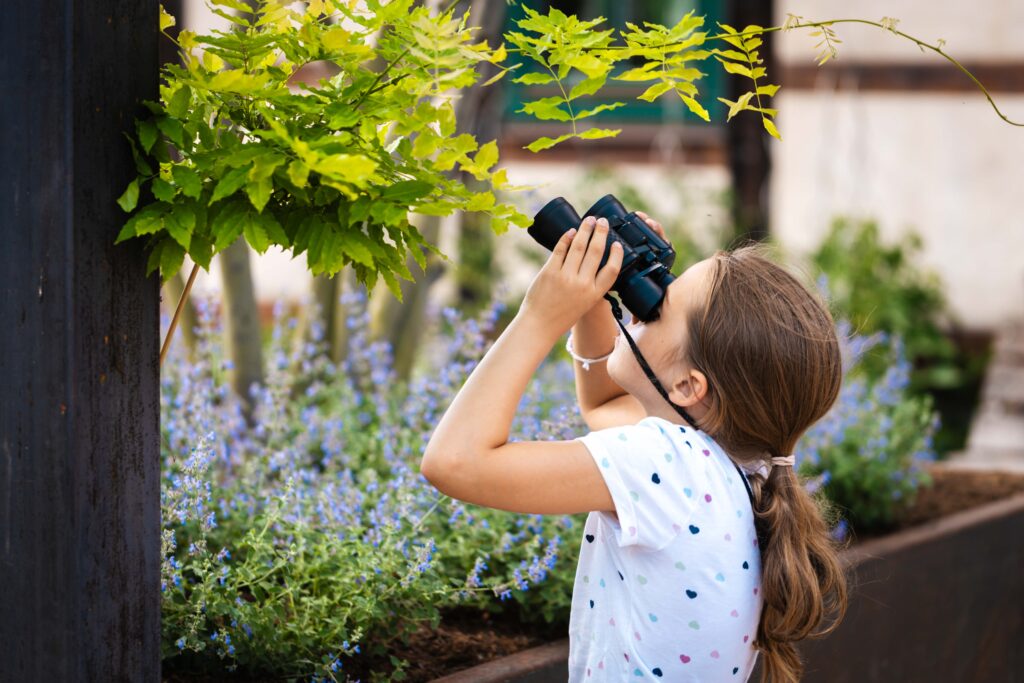 Mädchen hält Fernlas vor Augen und schaut im Garten vor Pflanze nach oben