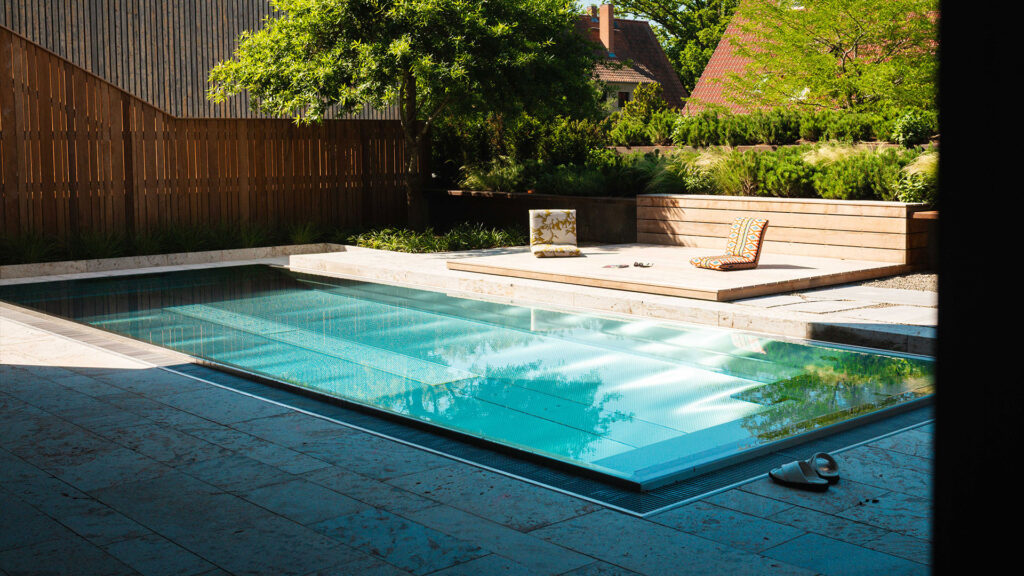 Ein moderner Außenpool mit kristallklarem, blauem Wasser, einer angrenzenden Holz-Sonnenterrasse mit Bodenkissen und üppiger Gartenbepflanzung im Hintergrund.