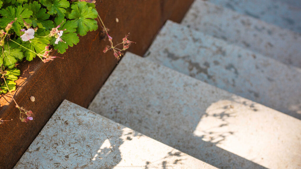 Detailaufnahme einer Treppe aus Naturstein neben einem Bepflanzungsbeet aud Cortenstahl