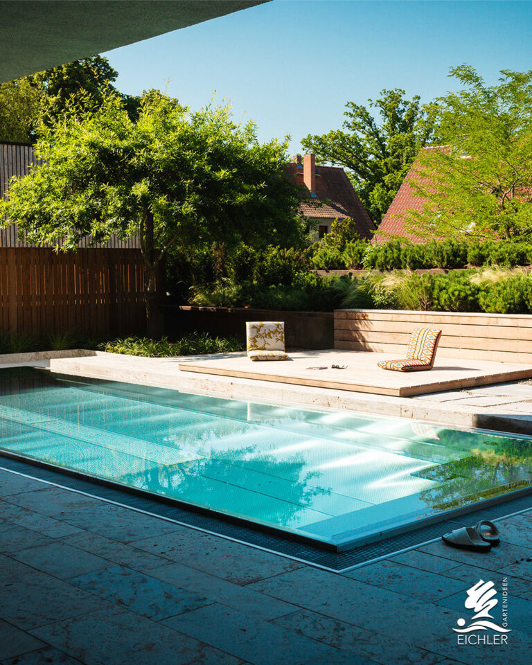 Ein moderner Außenpool mit kristallklarem, blauem Wasser, einer angrenzenden Holz-Sonnenterrasse mit Bodenkissen und üppiger Gartenbepflanzung im Hintergrund.