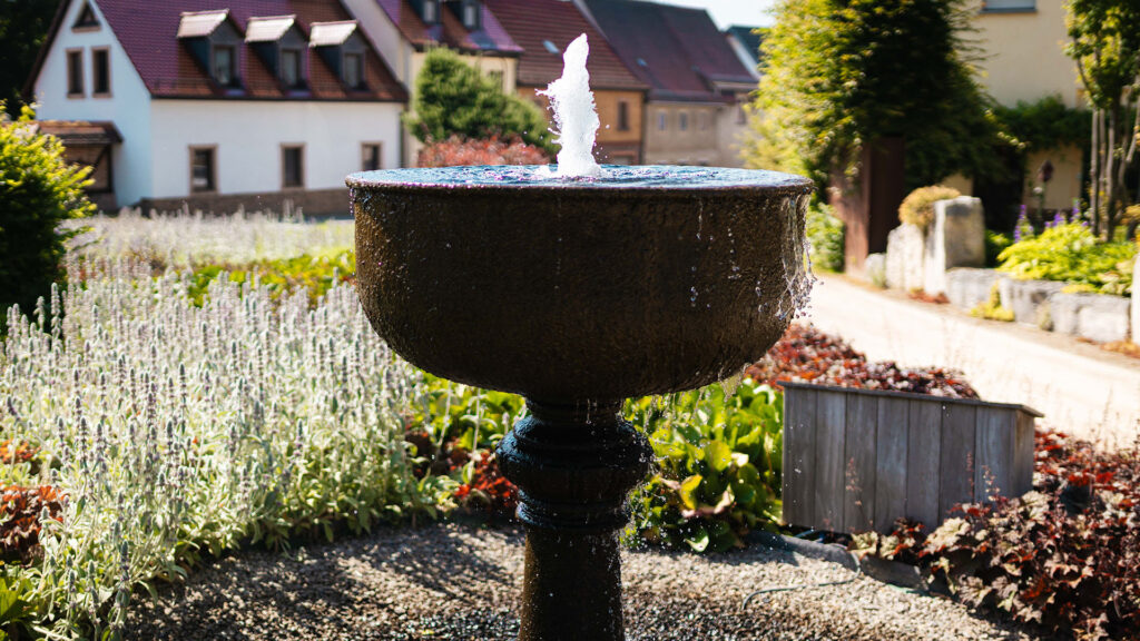 Klassischer kelchförmiger Gartenbrunnen mit Fontäne, eingebettet in ein Beet aus silbrigen Stauden und grünem Gebüsch.
