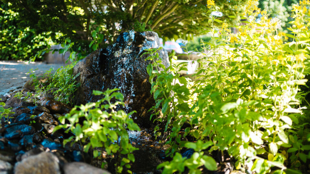 Ein dunkler Quellstein im Garten mit herunterfließendem Wasser, umringt von grünen Pflanzen und Flusskieseln.