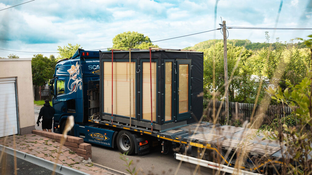 Blauer LKW-Tieflader befördert eine Sauna mit Glasfronten zur Baustelle.