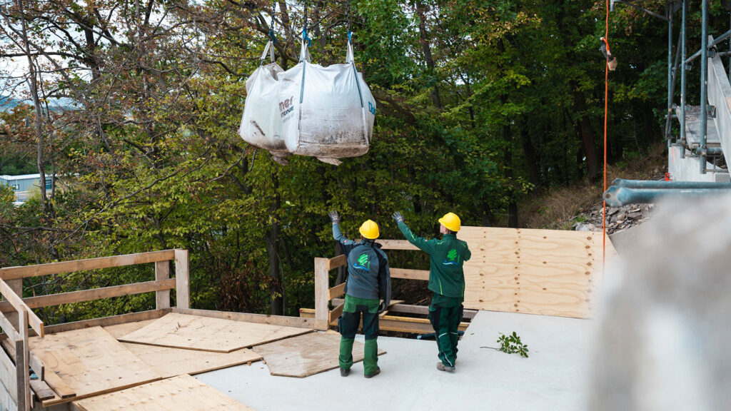 Zwei Bauarbeiter in Schutzbekleidung signalisieren und steuern die Ankunft von zwei großen Big Bags, die an einem Kran schweben.