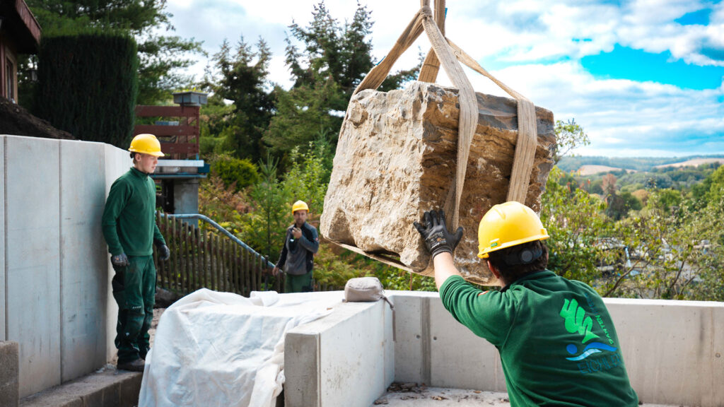 Zwei Gartenbauer mit gelben Schutzhelmen und grüner Arbeitskleidung führen einen großen, am Kran hängenden Naturstein auf einer Baustelle.