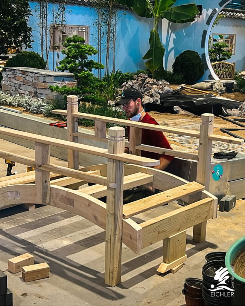 Ein Handwerker baut konzentriert an einer geschwungenen Holzbrücke in einer Schaugarten-Kulisse.